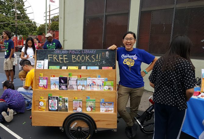 A person in a blue shirt stands next to a shelf of books attached to a bike.