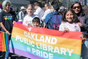 A group of young people stand behind a rainbow flag that says "oakland Public Library Pride"