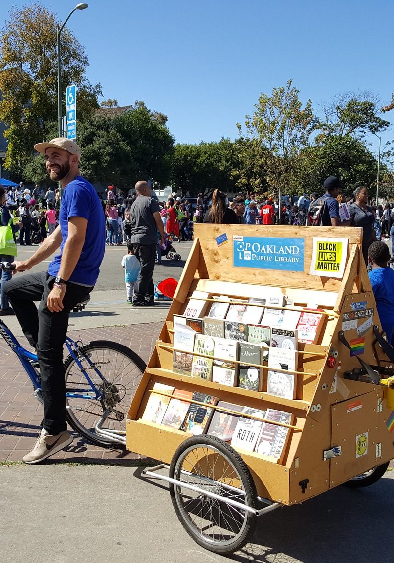 A man in a blue t-shirt sits on a bicycle attached to a shelf of books