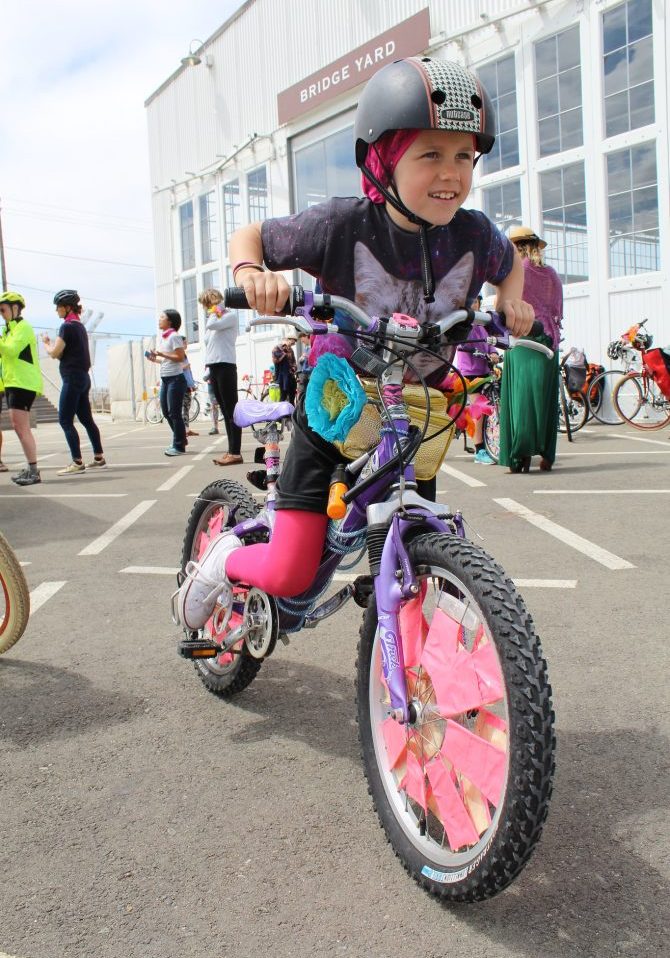 a smiling child, wearing a helmet and fuschia tights, rides a colorful scraper-style bicicyle